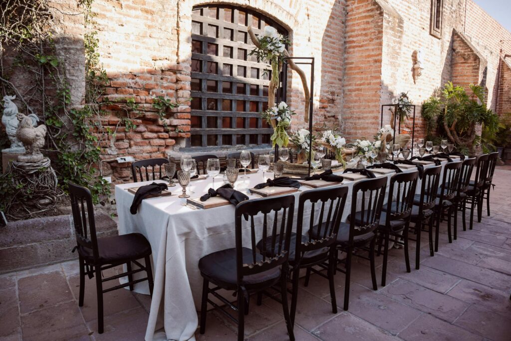 Elegant outdoor wedding table with floral decorations and black chairs against brick walls.